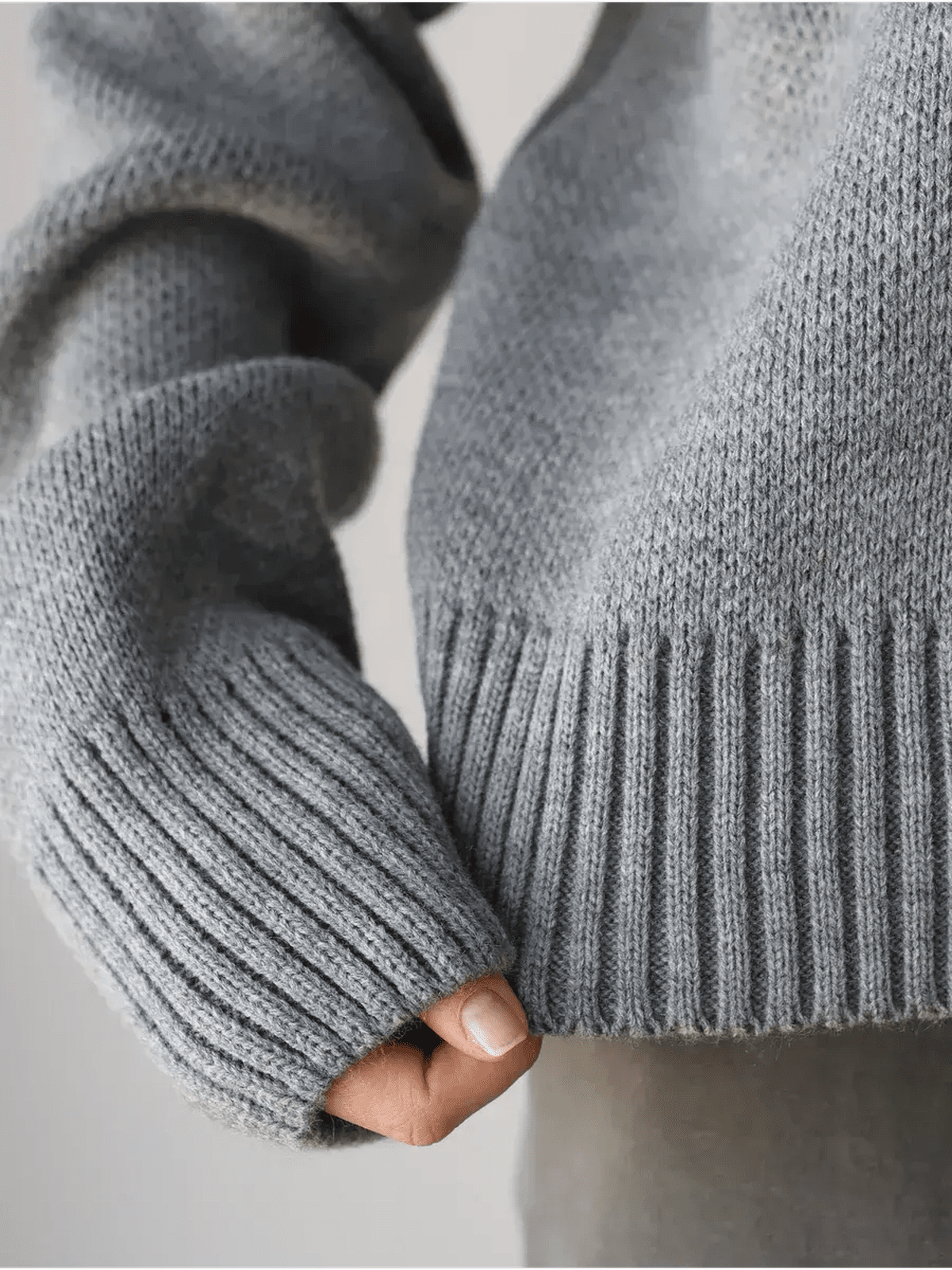 Close-up of a hand holding the hem of the Nova Pullover by Lauven, showcasing its soft gray knit and ribbed cuffs.