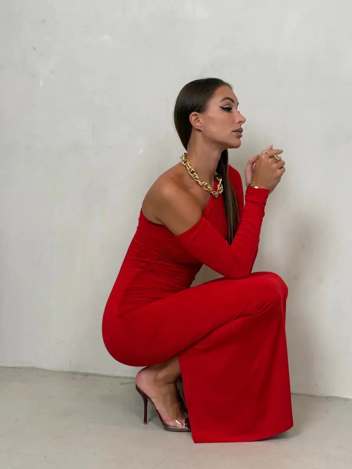 Elegant woman in a red one-shoulder maxi dress, showcasing a stylish crouch pose against a neutral background.