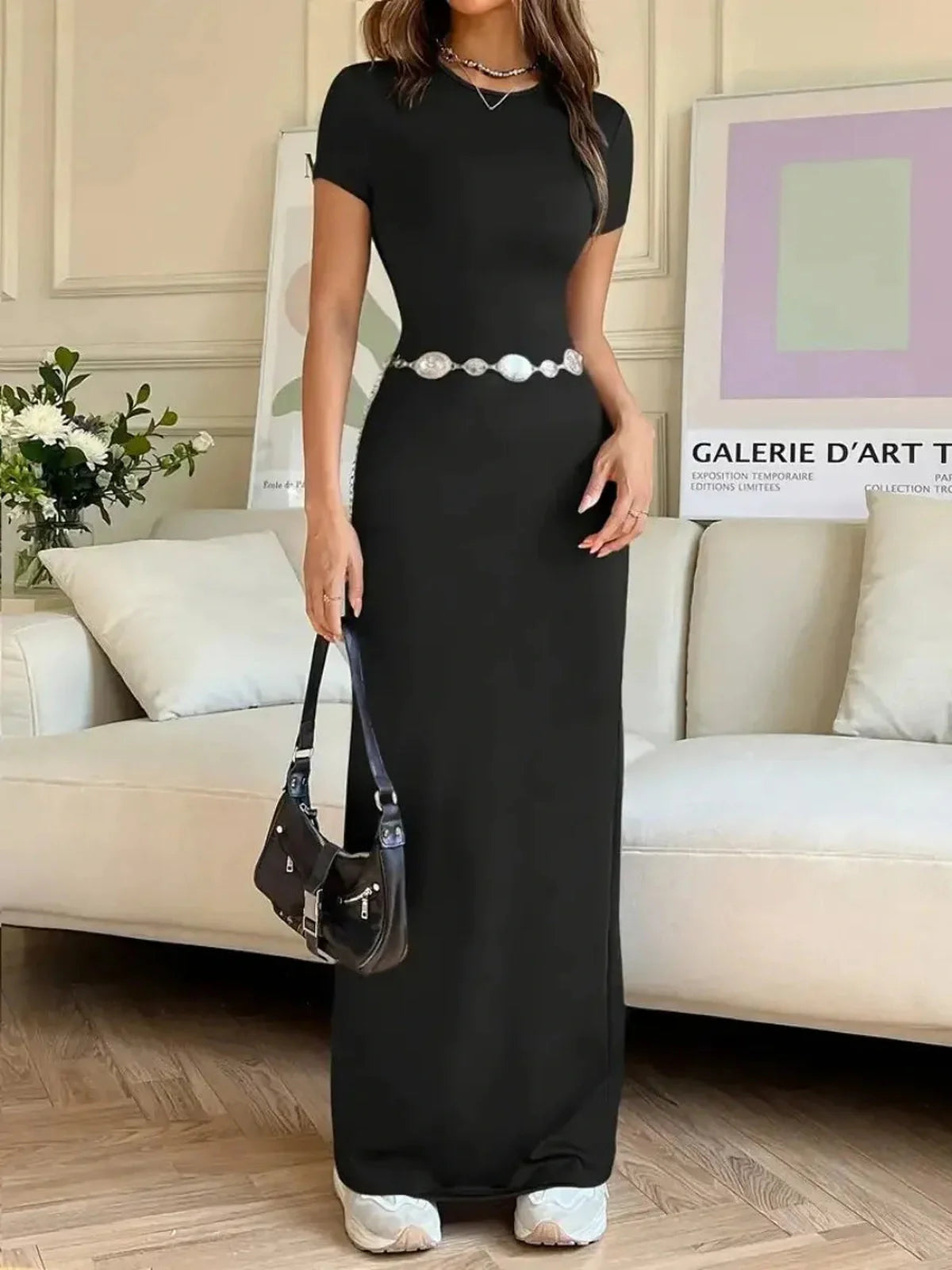 Model wearing a black Sculpted Grace Maxi Dress with a pearl belt, showcasing modern elegance in a stylish living room.