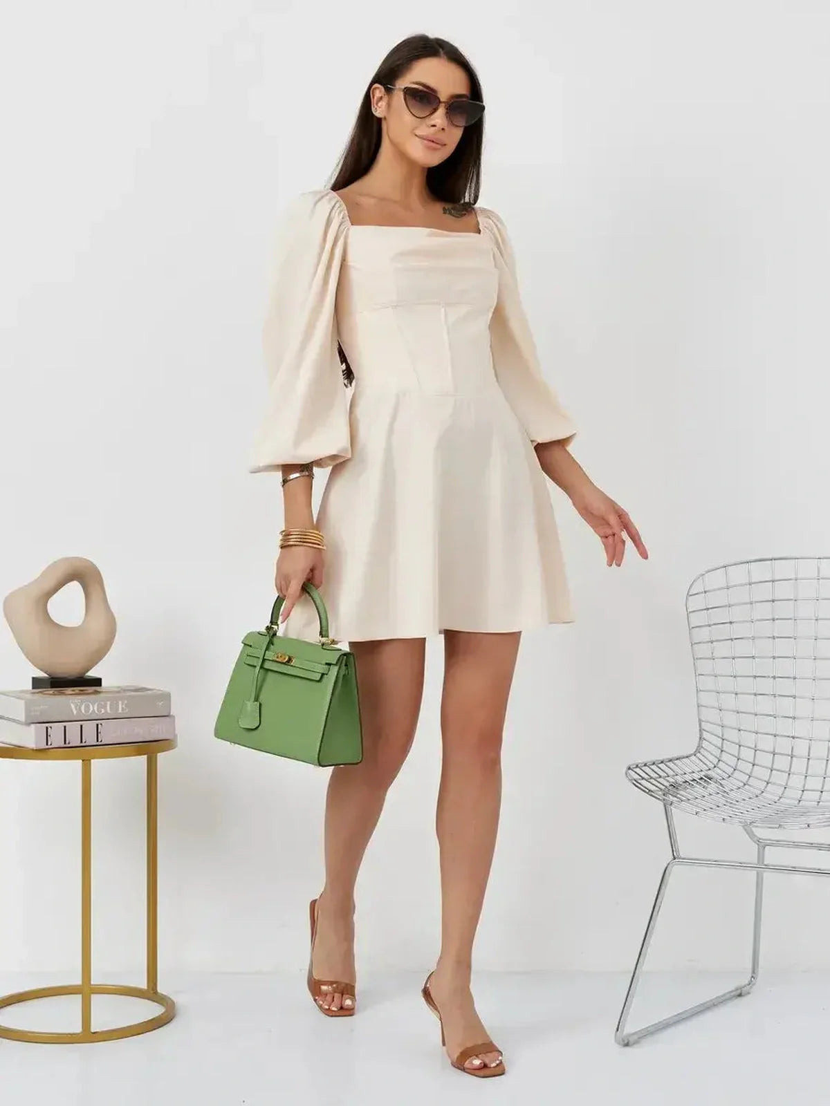 Model wearing Lace Muse beige mini dress with puff sleeves and square neckline, holding a green handbag.