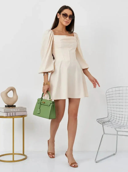 Model wearing Lace Muse beige mini dress with puff sleeves and square neckline, holding a green handbag.