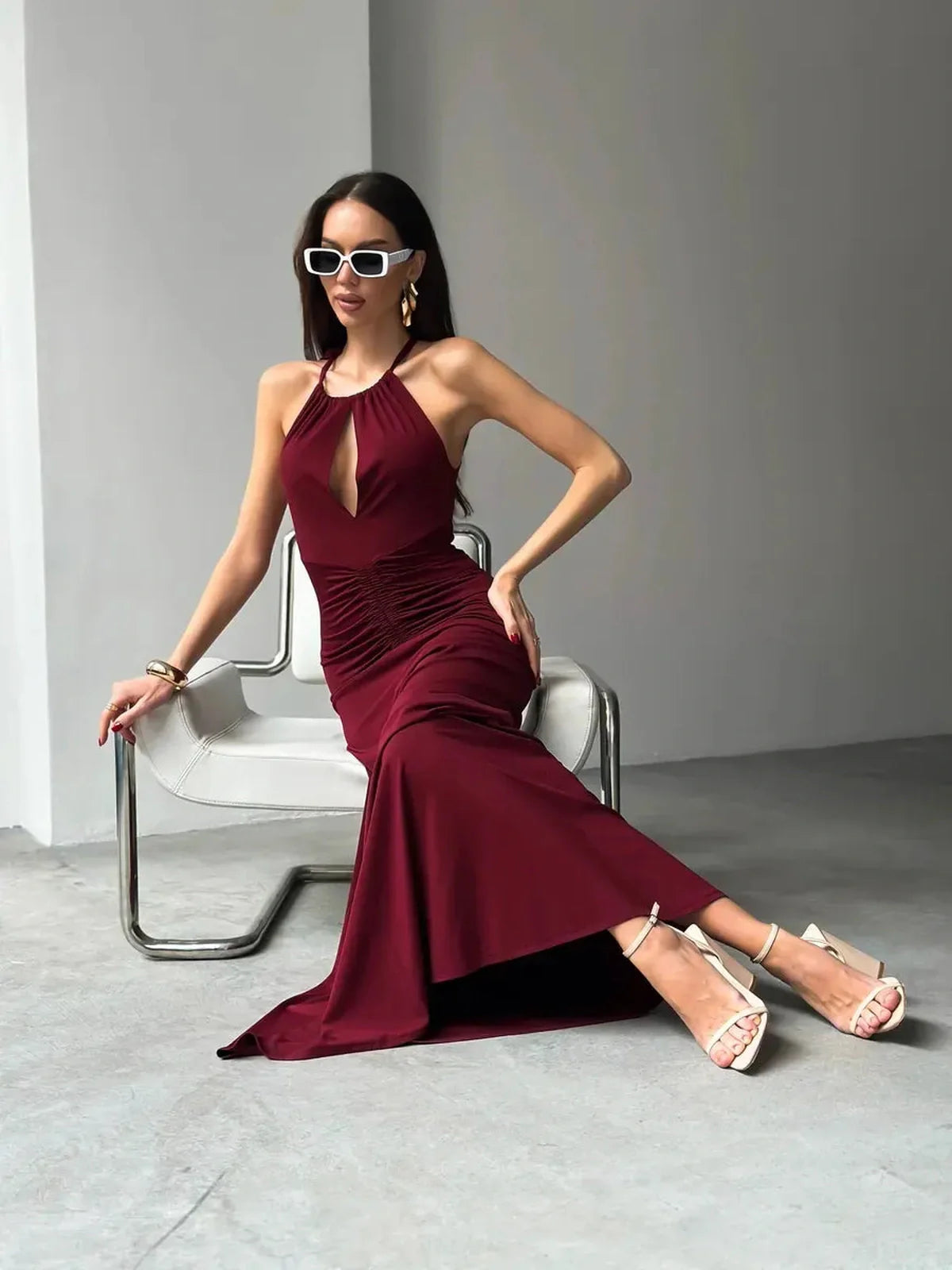 Model wearing a burgundy Maxi Dress Élan with draped halter neck, ruched waist, and stylish sunglasses, seated elegantly.