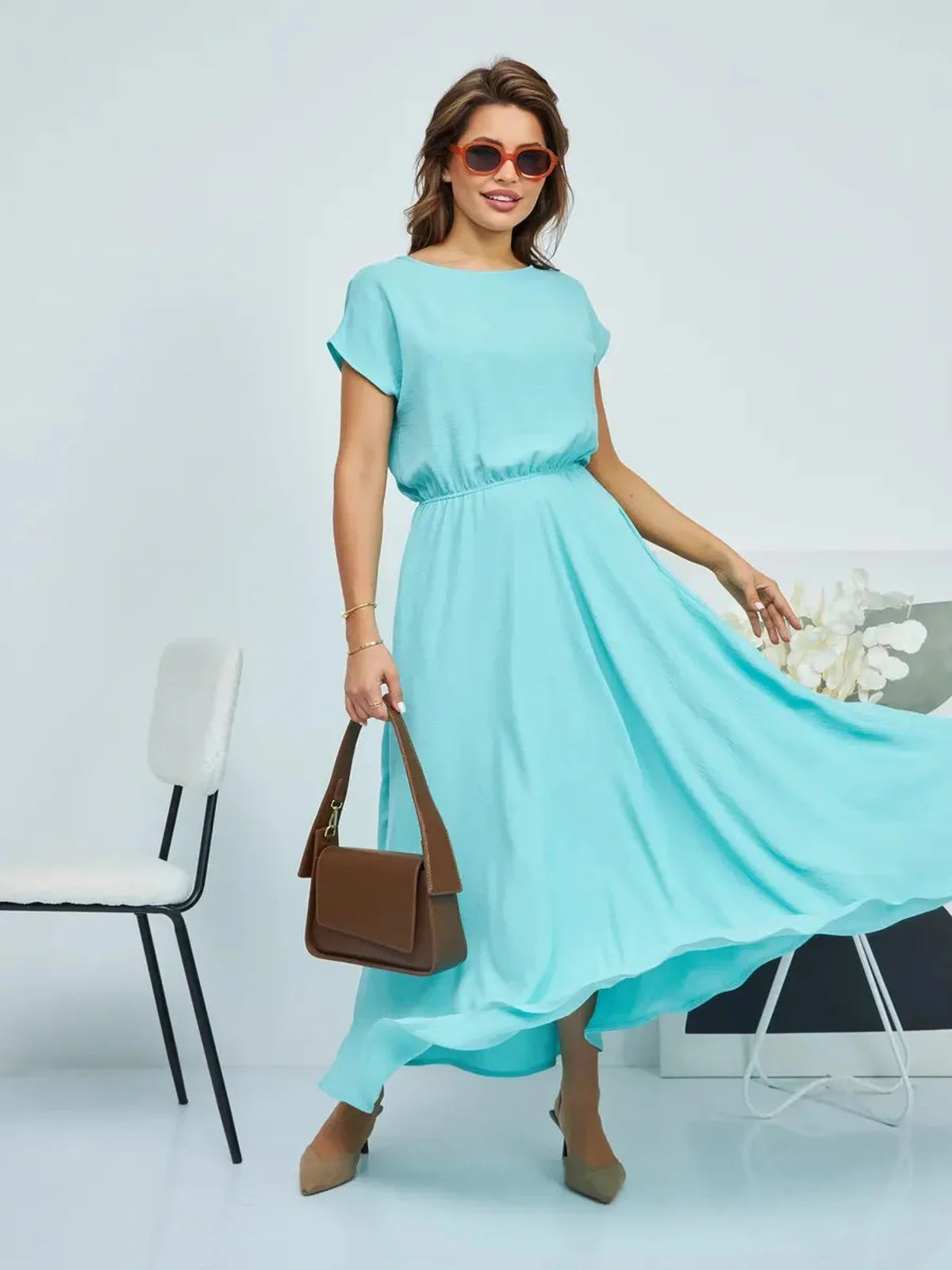 Model wearing a flowing aqua maxi dress with a belt, styled with sunglasses and a chic handbag.