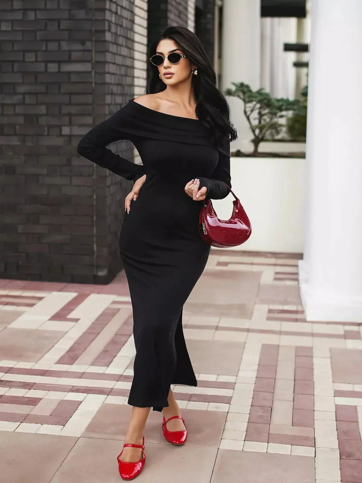 Woman in an iconic off-shoulder black maxi dress with a side slit, styled with red accessories and sunglasses.