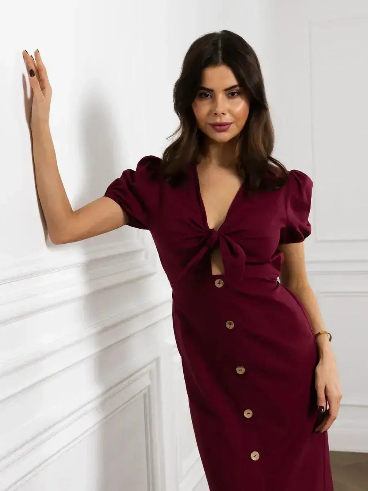Elegant woman in a burgundy maxi dress with front bow and button detail against a stylish white background.