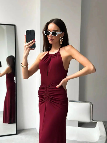 Model wearing a burgundy maxi dress with draped halter design and ruched waist, posing in front of a mirror.