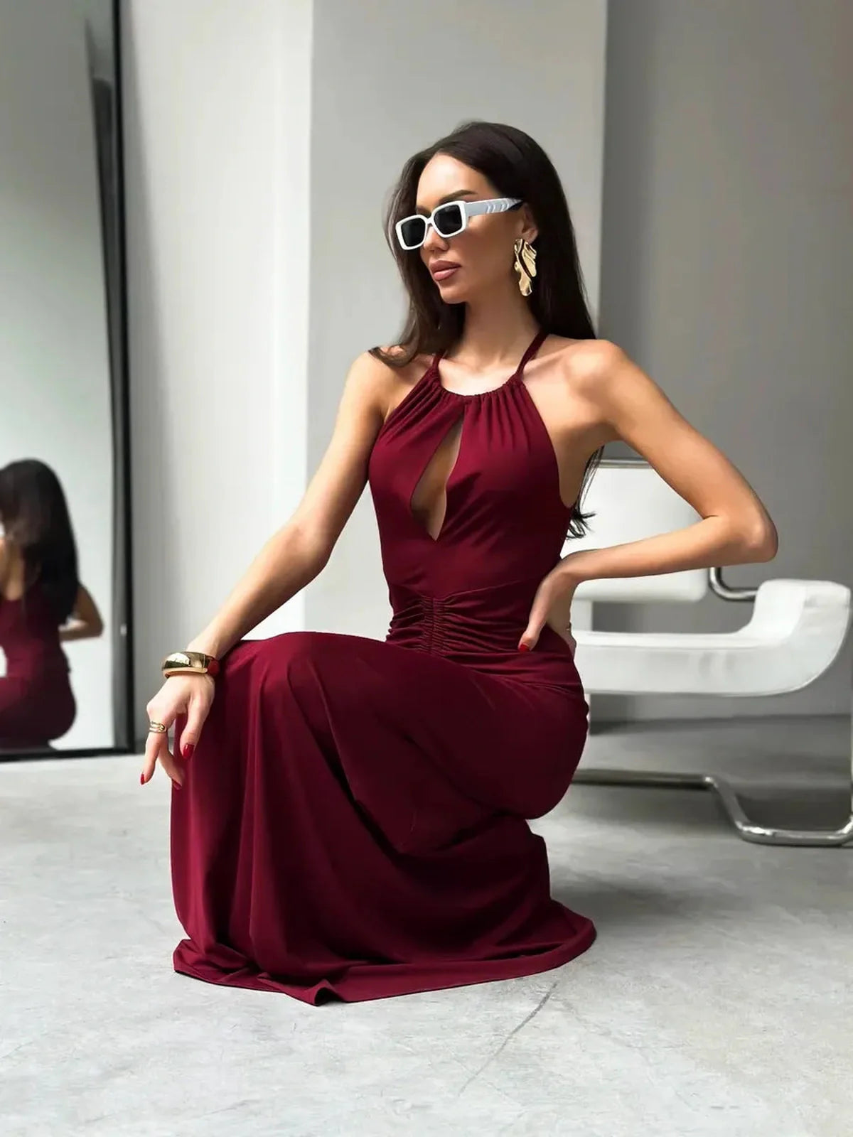 Elegant woman in a burgundy maxi dress with halter neckline and ruched waist, posing confidently in a modern setting.