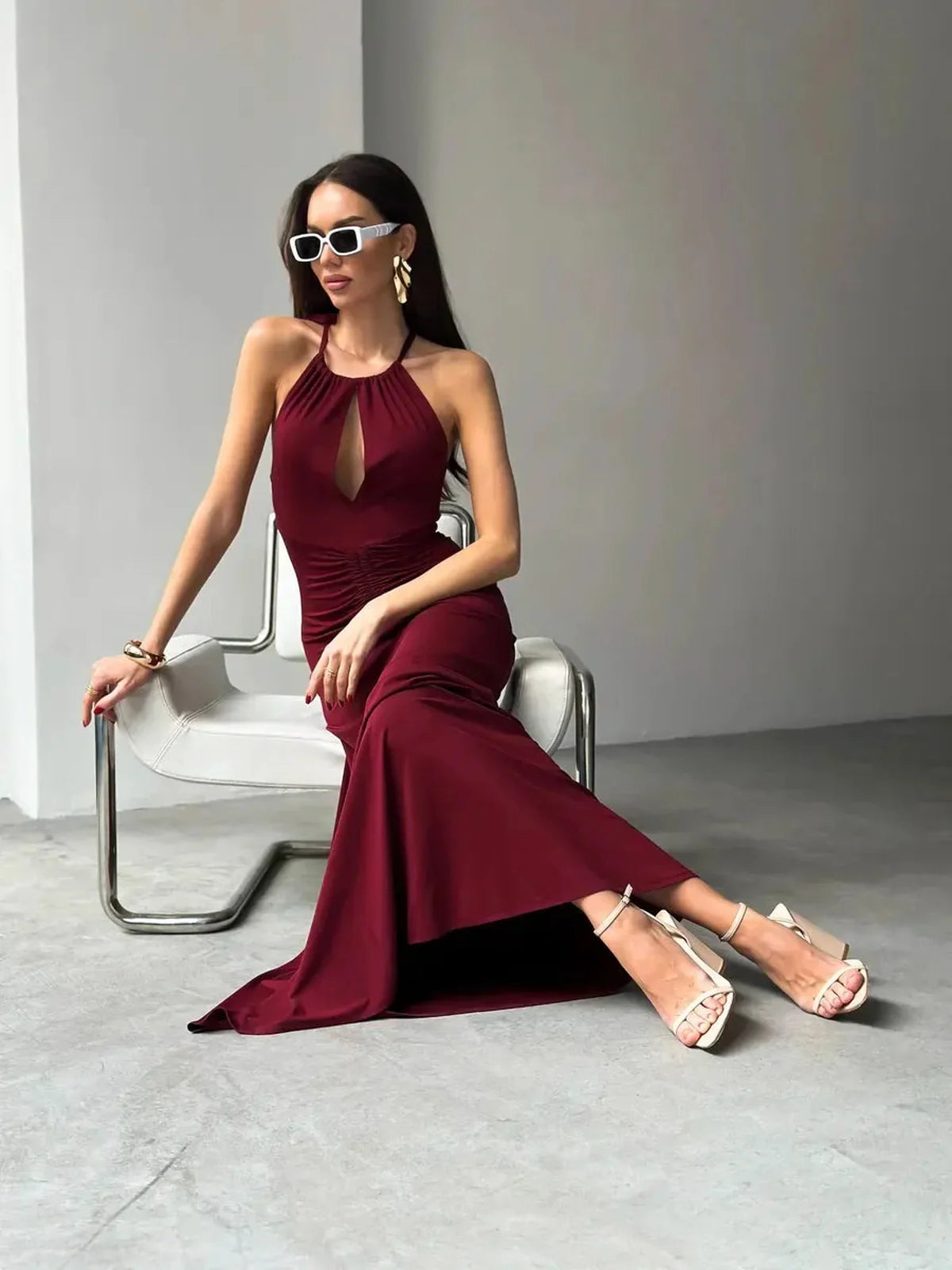Elegant woman in a burgundy maxi dress with draped halter design and front keyhole, styled with sunglasses and heels.