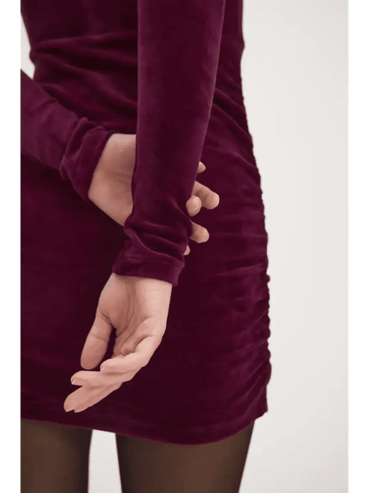 Close-up of a model's hand in a burgundy velvet mini dress with ruching detail, showcasing elegant long sleeves.