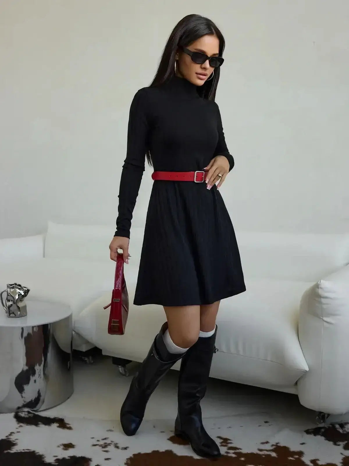 Elegant black ribbed mini dress with a high collar, styled with a red belt and knee-high boots.