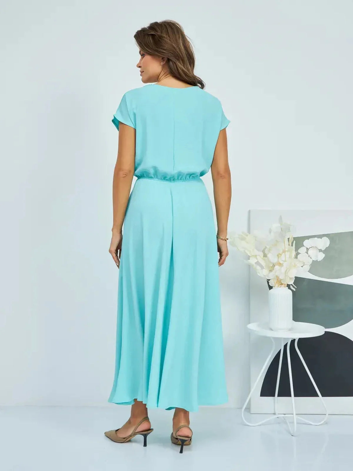 Woman in a light teal maxi dress showcasing a draped back design, styled with elegant shoes and a modern decor.