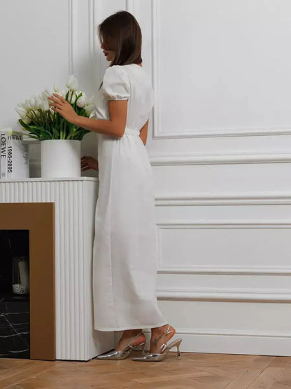 Woman in white maxi dress with puff sleeves, standing beside a plant, showcasing minimalist elegance.