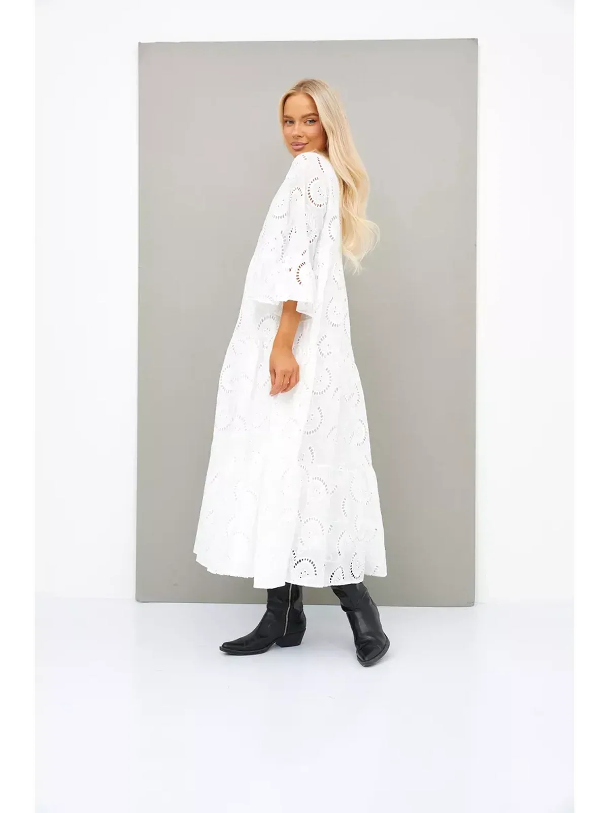 Elegant woman wearing a white broderie cotton maxi dress with flared sleeves and V-neck, styled with black boots.