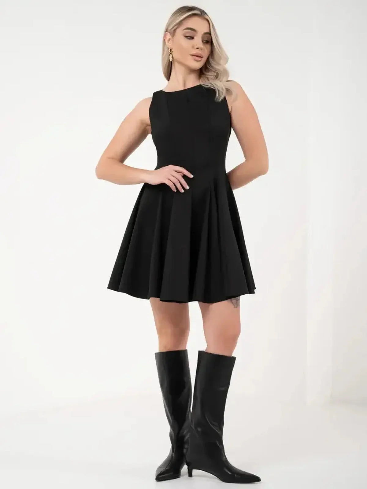 A model wearing a sleeveless black dress with a flared skirt, paired with knee-high black boots.