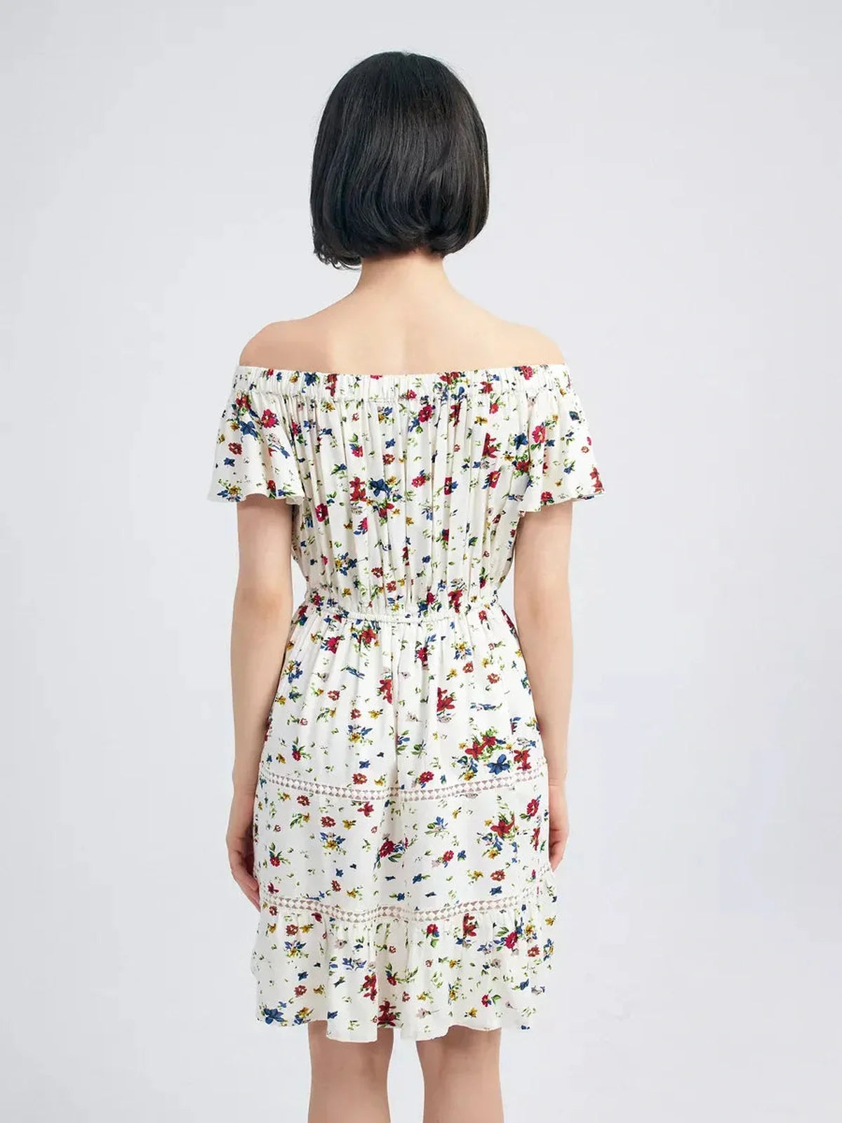 Back view of a woman in a white Bloom Off-Shoulder summer dress featuring multicolor floral print and embroidered trims.