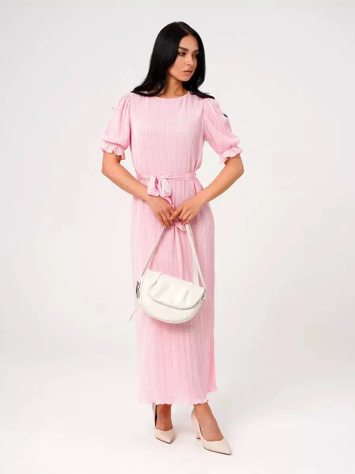 Model wearing a pink pleated Celeste Maxi Dress with puff sleeves and a tie waist, holding a white handbag.