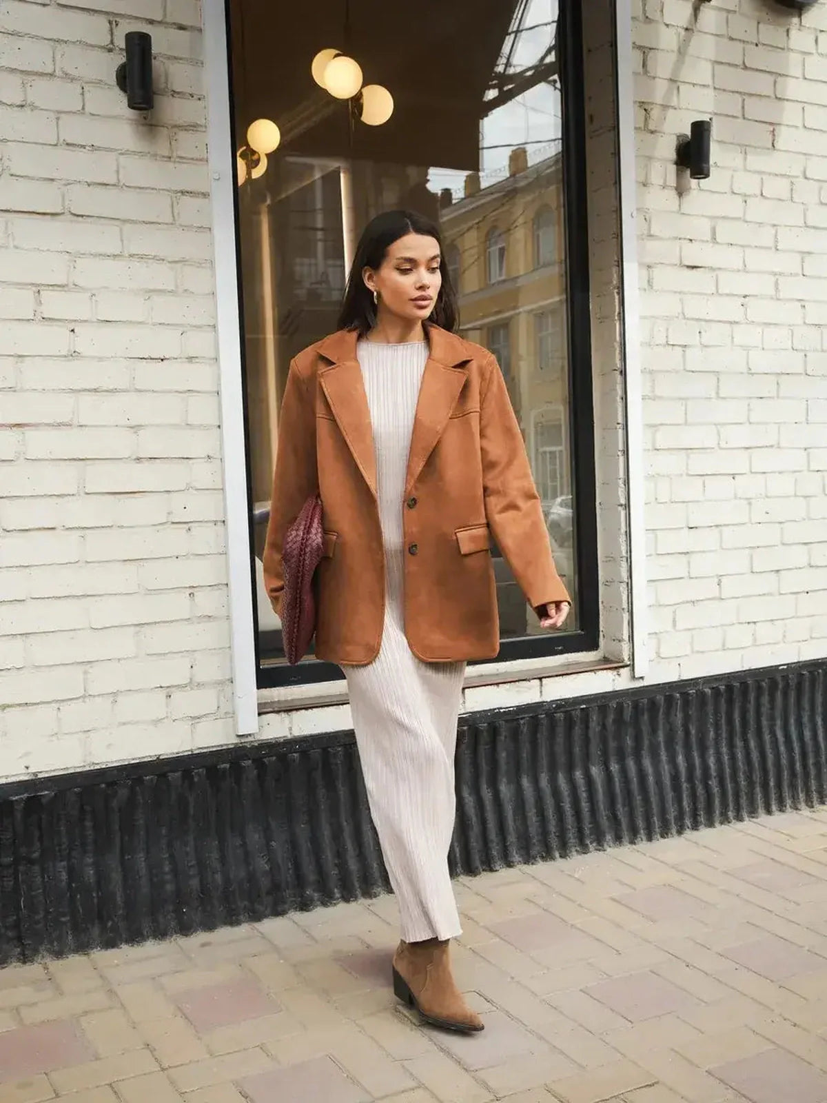 Stylish woman in a brown blazer over a cream contour knit maxi dress, walking by a storefront.
