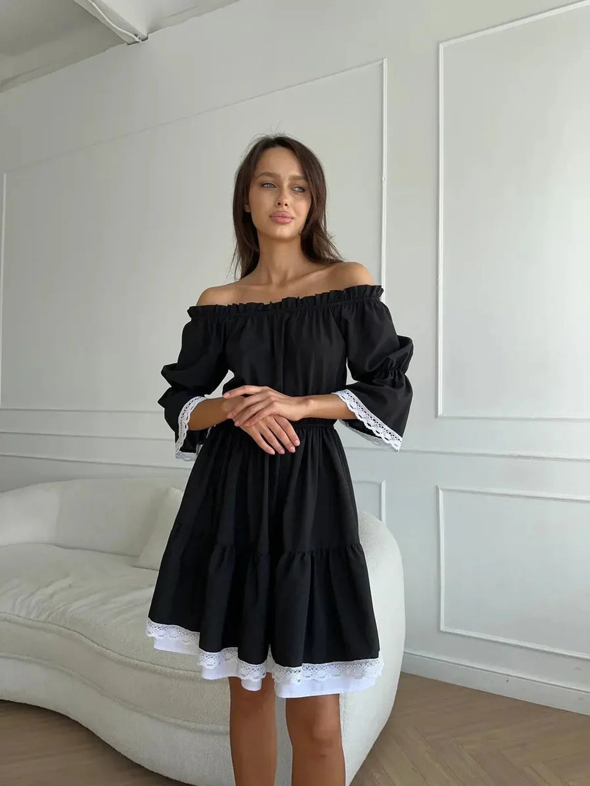 Woman wearing a black off-shoulder mini dress with white lace trim, posing in a stylish indoor setting.