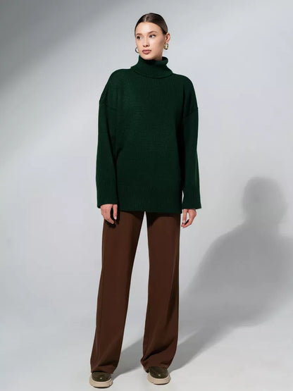 Model wearing Lyra Sweater by Lauven, an oversized green knitwear with turtleneck, paired with brown trousers.