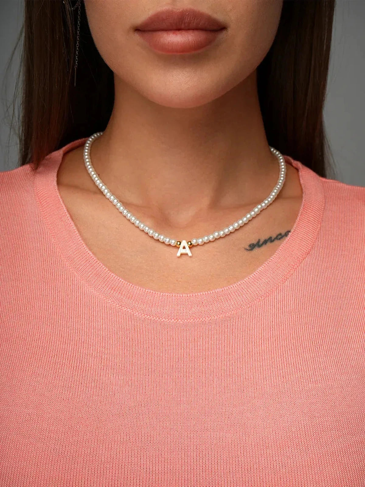 Woman wearing a pearl necklace with an initial 'A' on a soft pink shirt, showcasing modern elegance.