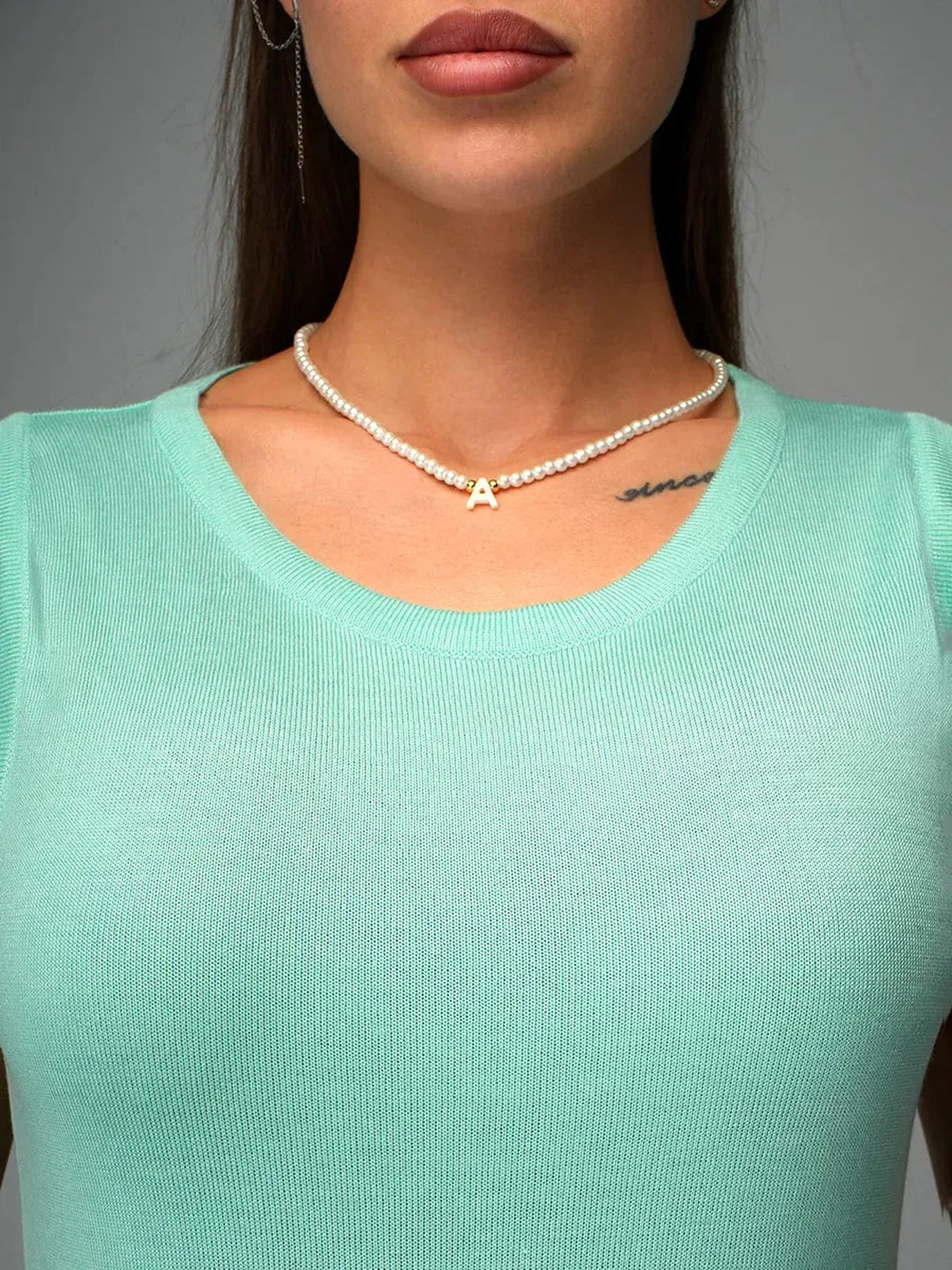 Close-up of a woman wearing a mint top, showcasing a pearl necklace with a gold letter 'A' charm.