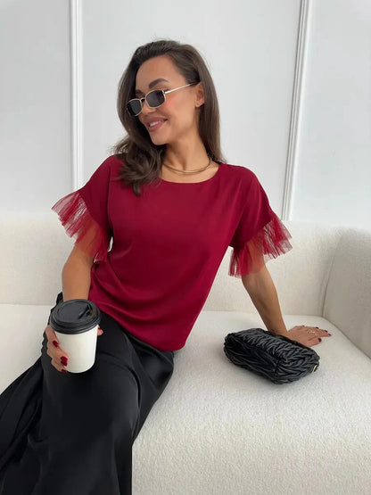 Woman in burgundy Velora Blouse with sheer ruffle sleeves, holding a coffee cup, exuding elegance and style.