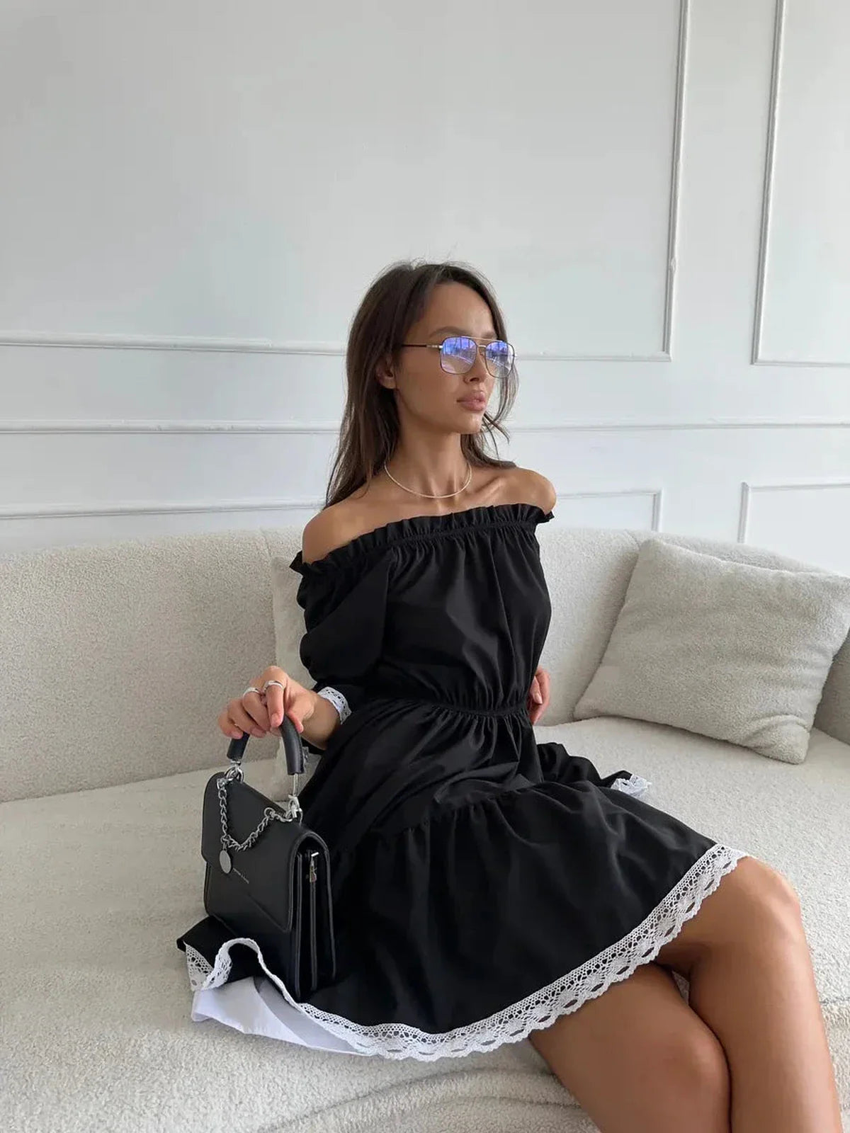 Model wearing the Contrast Charm black off-shoulder mini dress with white lace trim, styled with sunglasses and a handbag.