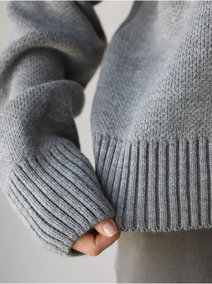 Close-up of a hand holding the hem of the Nova Pullover by Lauven, showcasing its soft gray knit and ribbed cuffs.