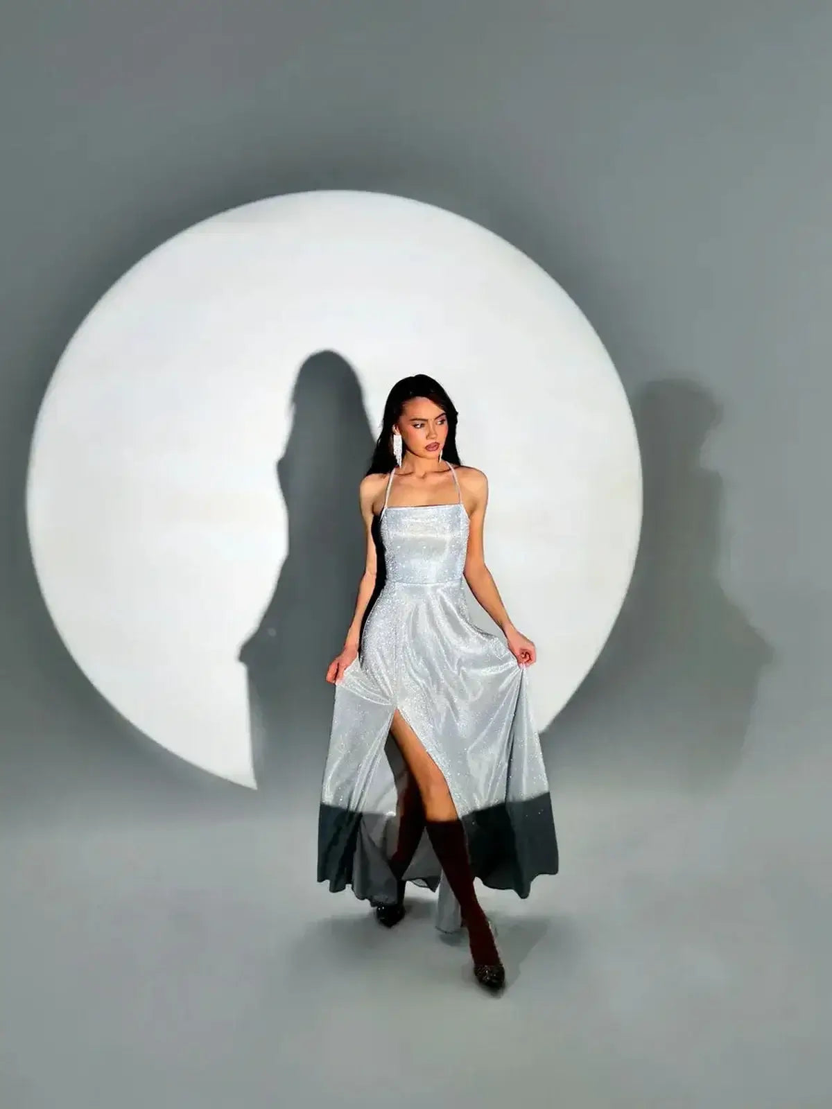 Model showcasing the Lunar Halo Maxi Dress, a silver gown with a side slit, against a dramatic circular backdrop.