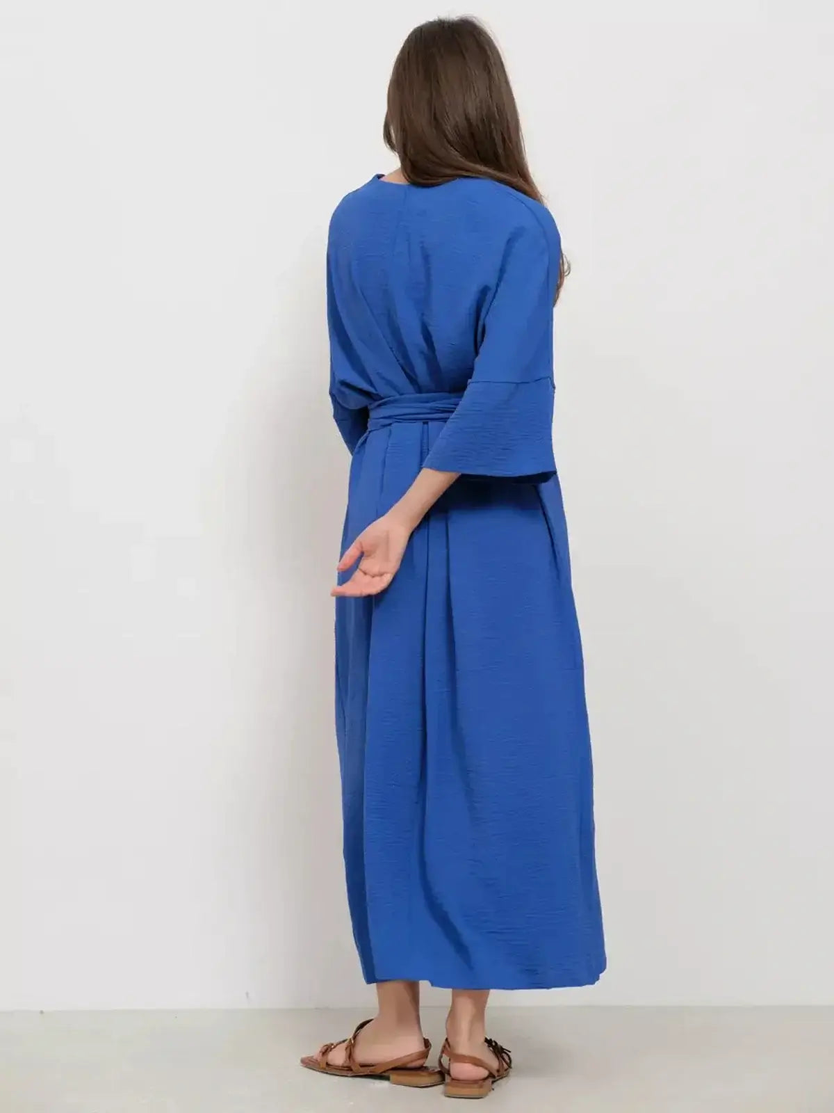 Woman wearing a long blue maxi dress with a relaxed fit and minimalist tie-belt from the back.