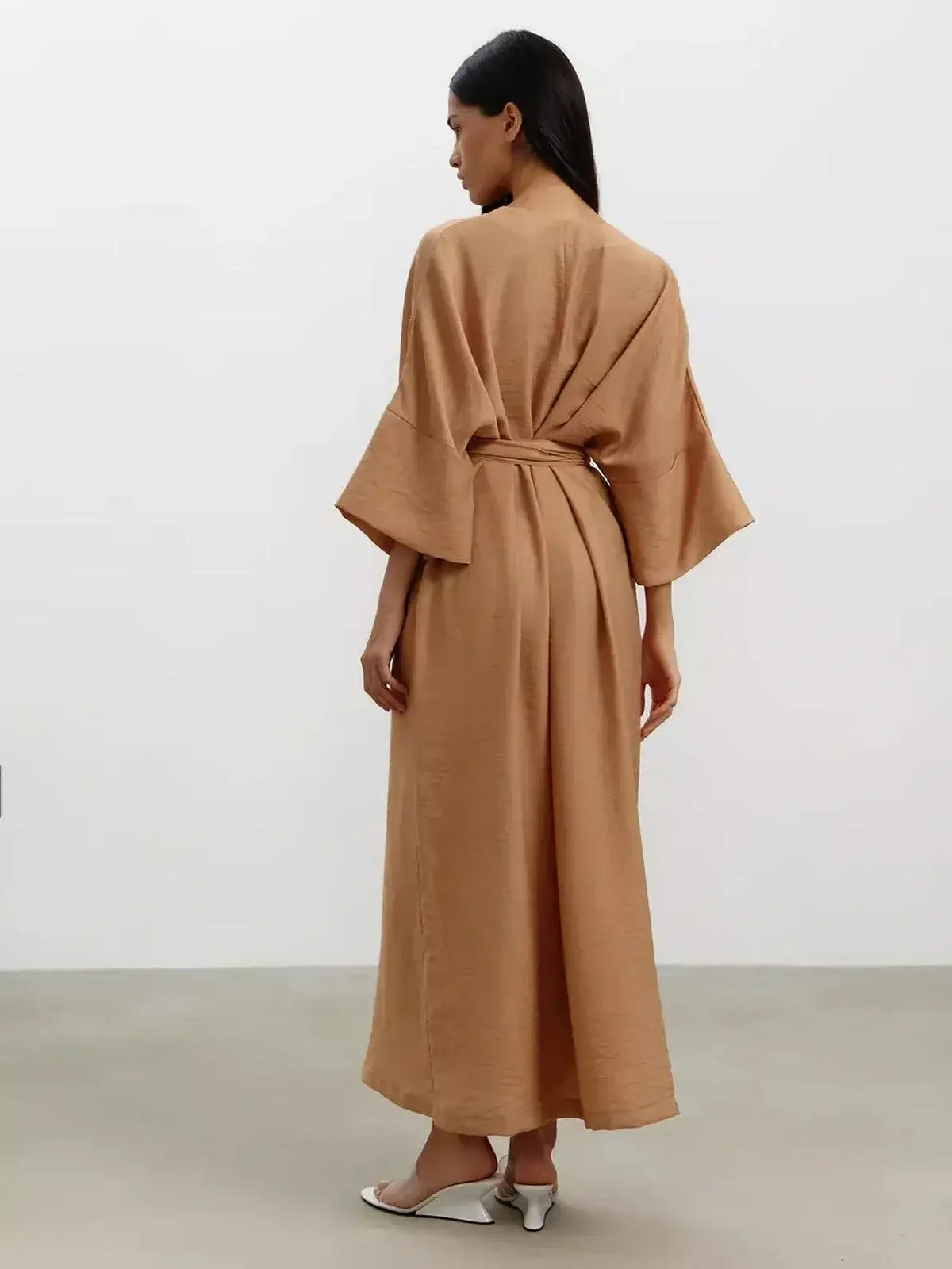 A woman in a flowing beige maxi dress with wide kimono sleeves and a tied waist, showcasing minimalist elegance.