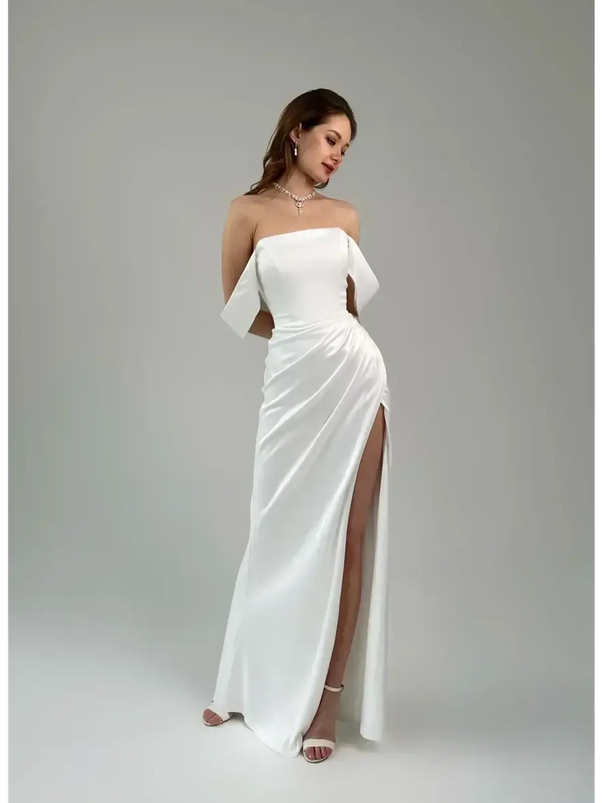 Elegant woman in off-shoulder white satin maxi dress with thigh-high slit and draped design.