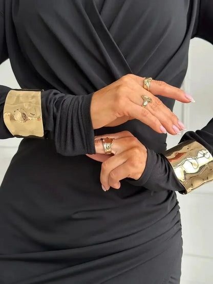 Close-up of hands showcasing stylish rings and elegant gold cuffs on black long-sleeve dress, highlighting modern fashion details.