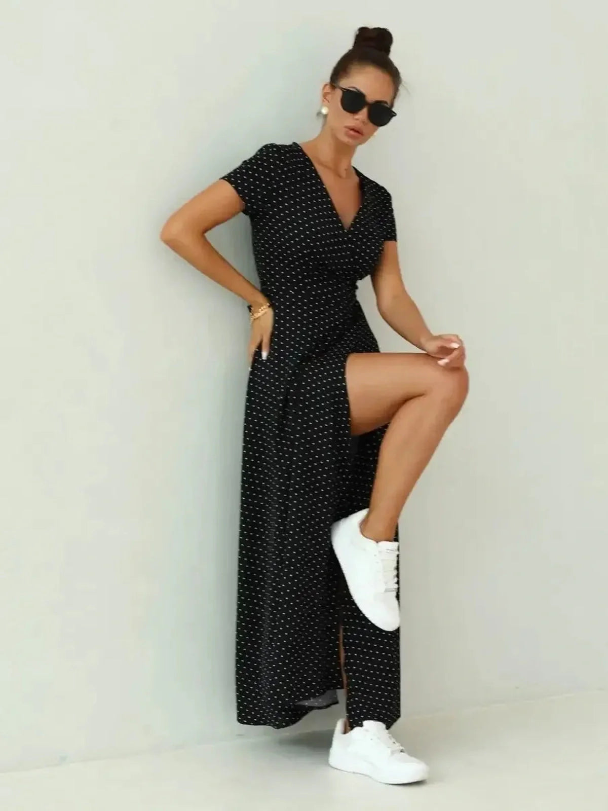 Model wearing a black polka dot maxi dress with a wrap silhouette and thigh-high slit, paired with white sneakers.
