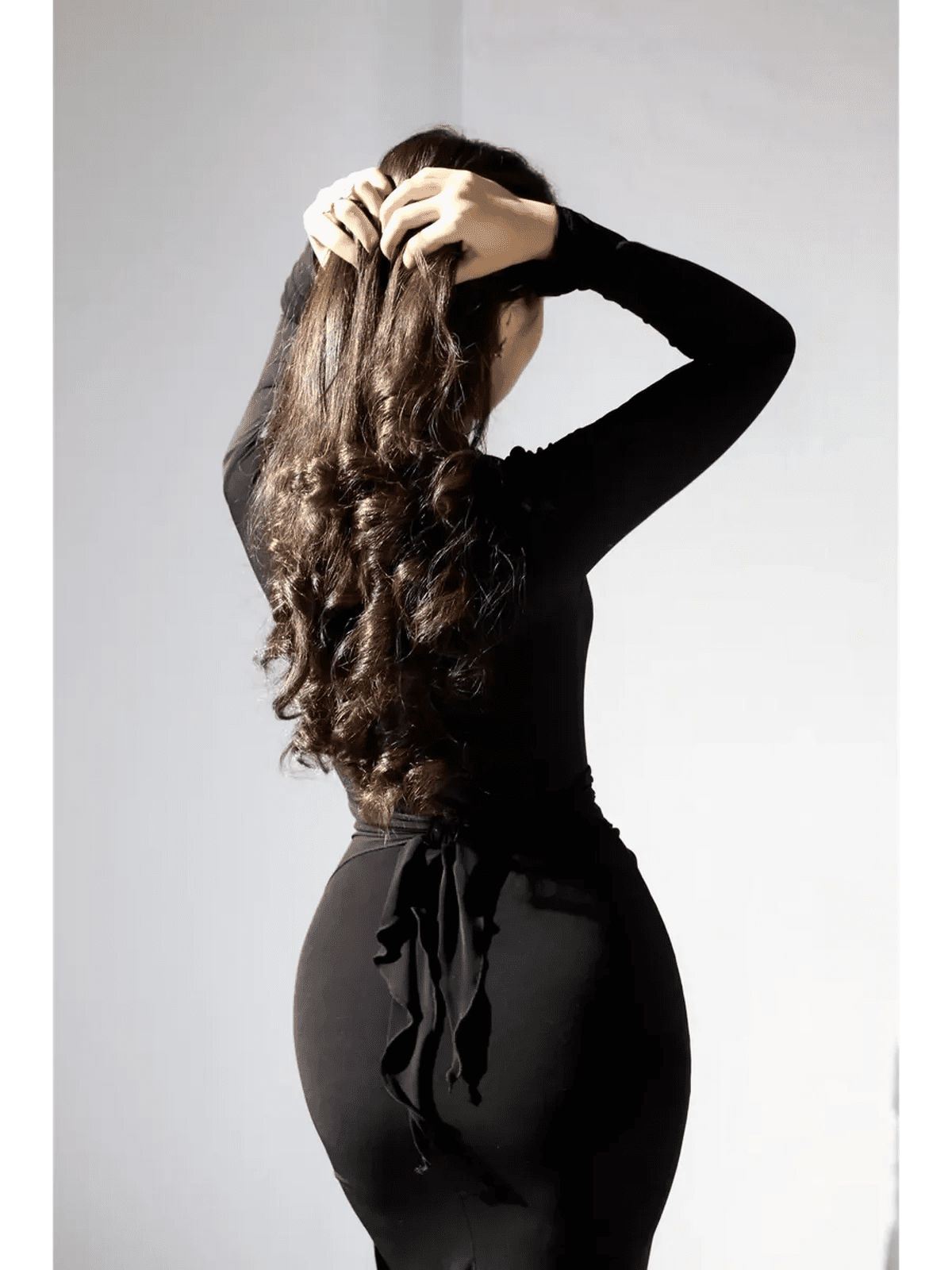 Elegant woman adjusting her hair while wearing a sleek black maxi dress with a tie detail at the back.