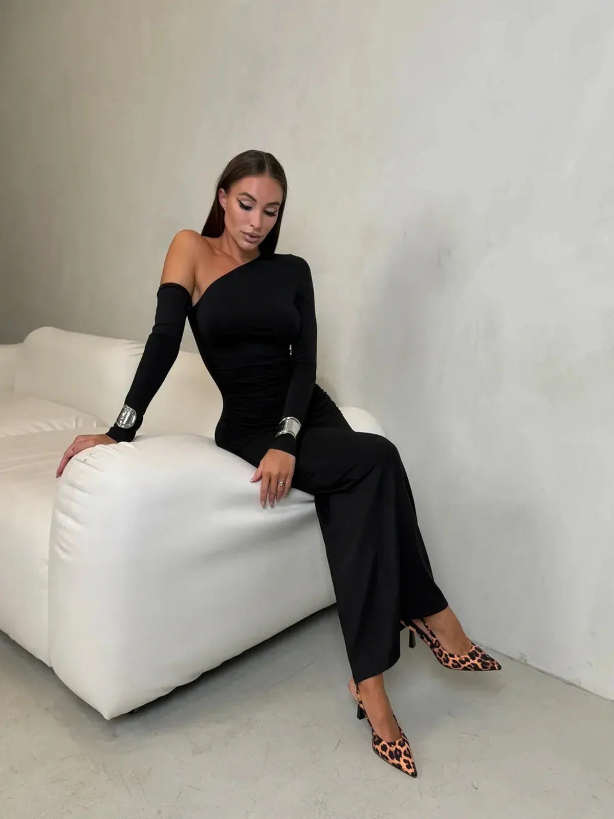 Model showcasing the Obsidian Maxi Dress with an asymmetric neckline, seated elegantly on a white couch.