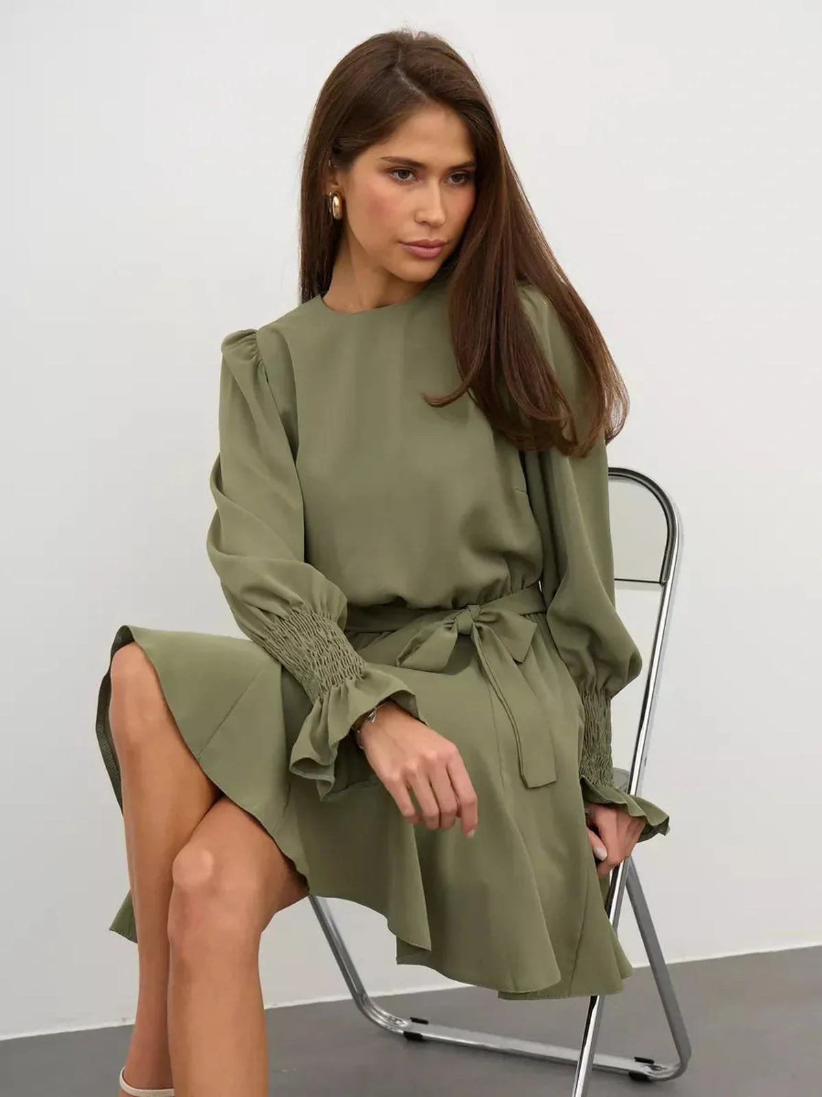 Model wearing the Olive Luxe Mini Dress in olive green, featuring long sleeves, ruffle details, and a waist-sash.