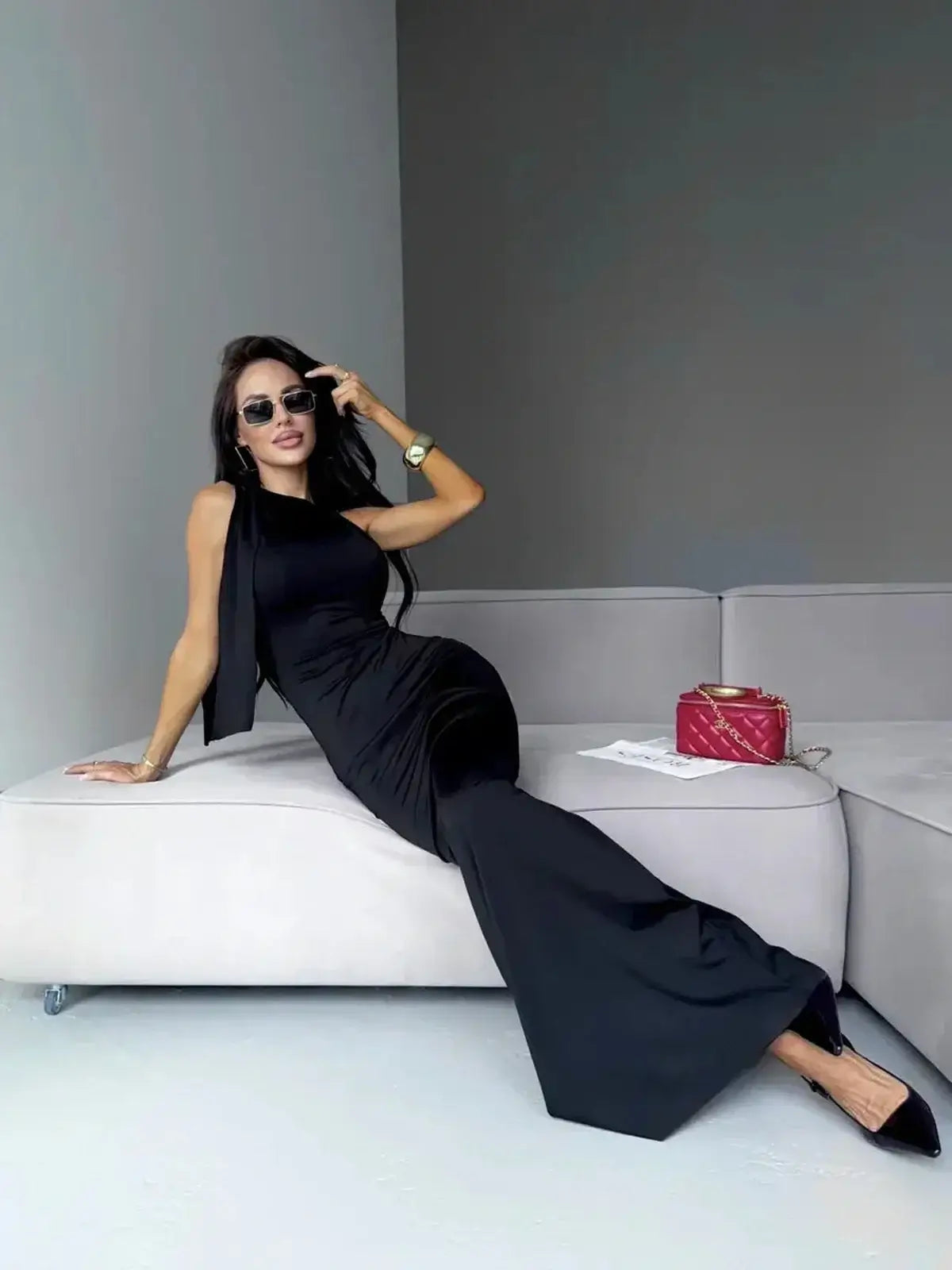 Model wearing a sleek black Sculpted Noir Maxi Dress, showcasing an elegant bow detail while relaxing on a stylish sofa.