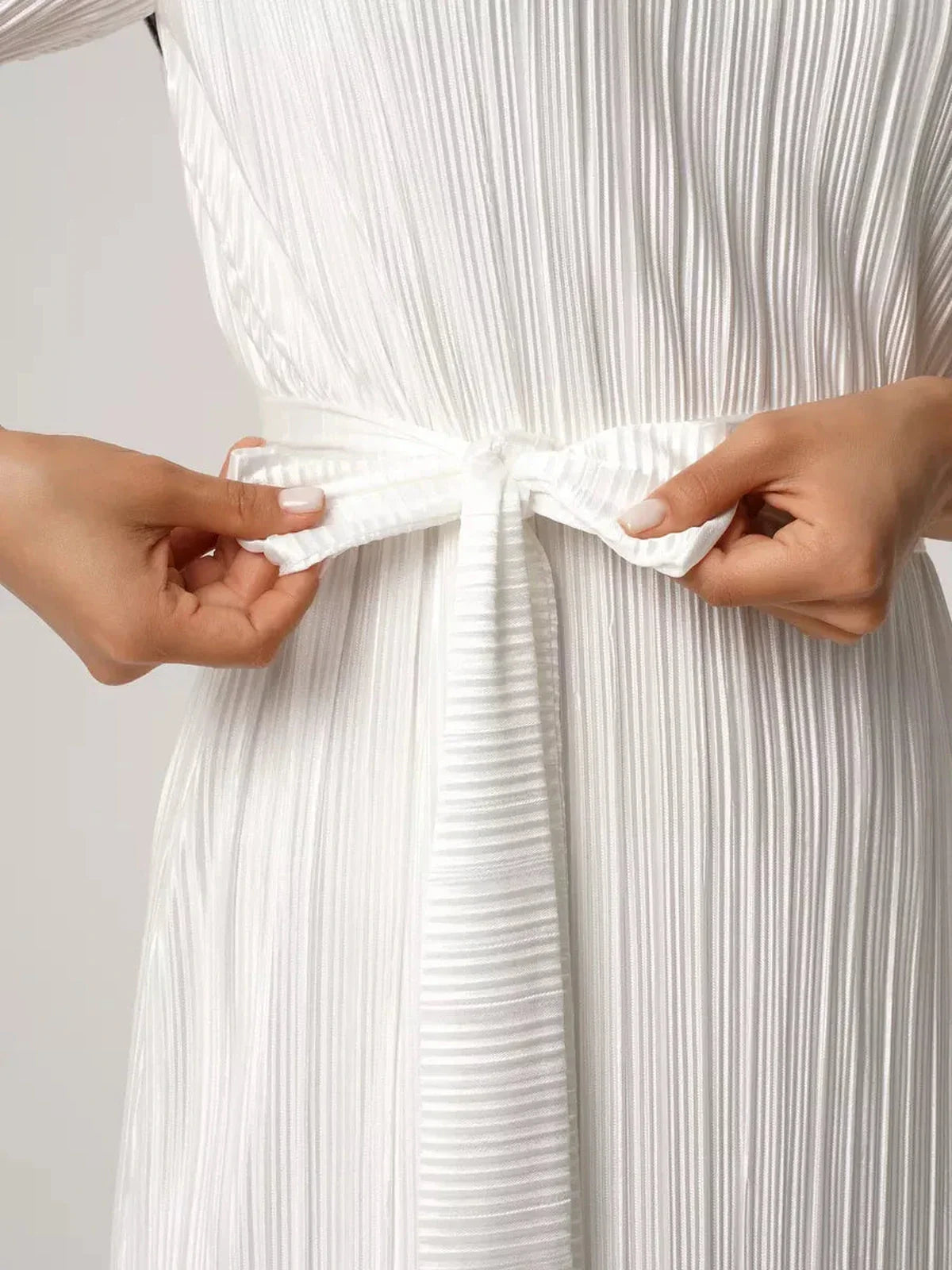 Model adjusting the bow tie of a pleated Seraphine Maxi Dress, showcasing its elegant fabric texture in white.