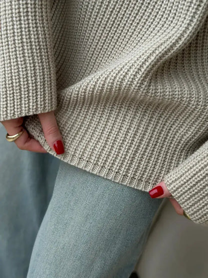 Close-up of hands holding the hem of the oversized Sierra Sweater by Lauven, showcasing its beige knit texture.