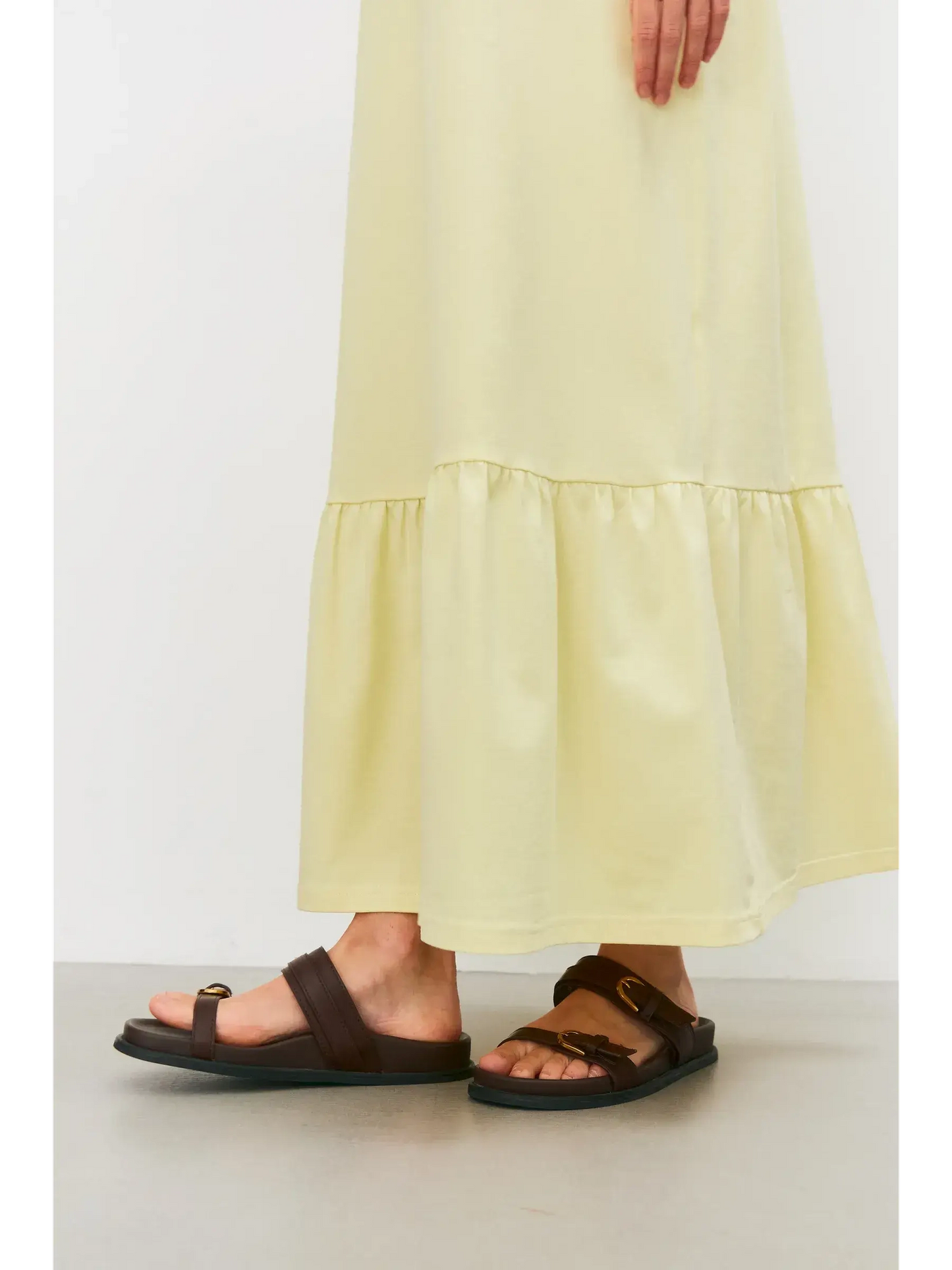 Close-up of sandals paired with a light lemon maxi dress showcasing its gathered hem flounce for a casual summer look.