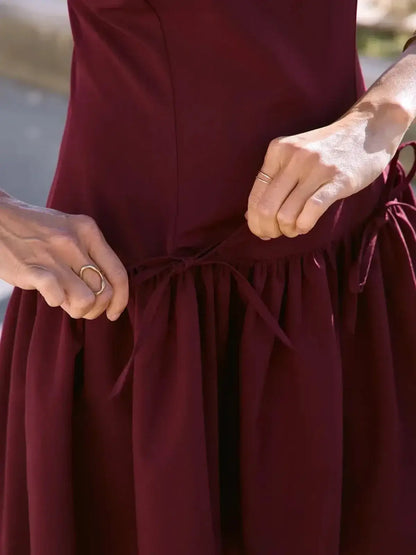 Close-up of hands tying the bow on a burgundy dress, showcasing its elegant design and pleated skirt.
