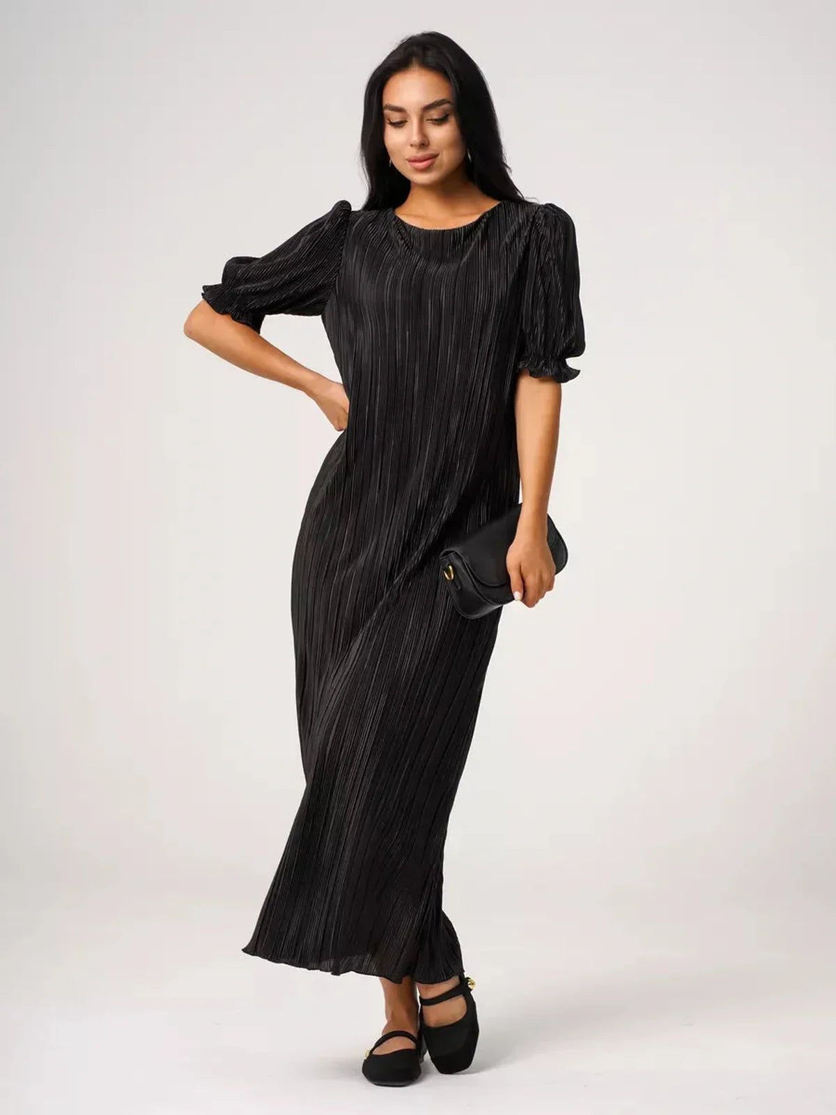 Elegant woman in a pleated black Velvet Rhythm Maxi Dress with puff sleeves, holding a clutch.