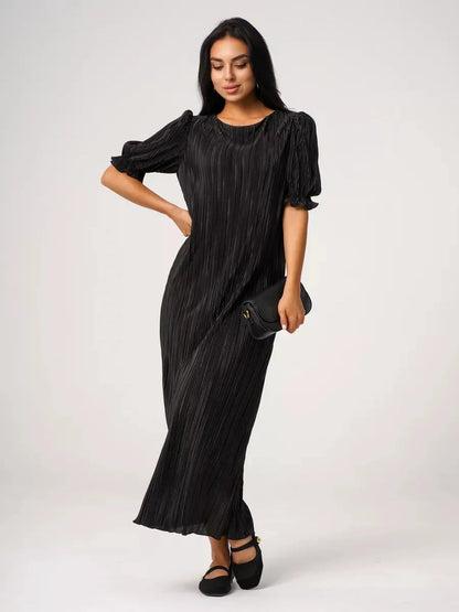Elegant woman in a pleated black Velvet Rhythm Maxi Dress with puff sleeves, holding a clutch.